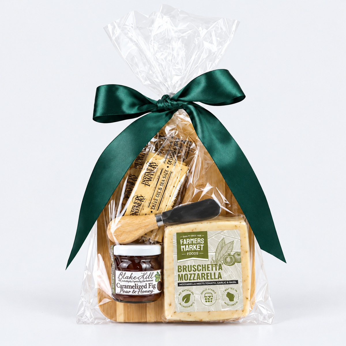 A gourmet gift hamper from Farmers Market Gourmet, wrapped in clear cellophane and tied with a dark green satin ribbon. The package contains a block of Bruschetta Mozzarella cheese, a jar of caramelized fig jam, a small knife, and packaged crackers on a wooden board, set against a clean white background.